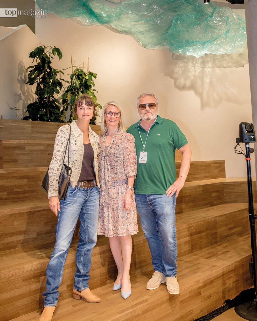 Giulia Poppi (l.) mit Geschäftsführerin Theresa von Fugler (Sanofi Consumer Healthcare) und Künstler Tobias Rehberger unter ihrem Kunstwerk aus Plastik, das wie Wasser aussieht