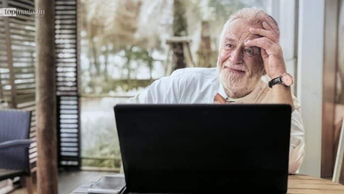 Ein stilvoller Senior mit weißem Bart sitzt nachdenklich vor seinem Laptop und nimmt am digitalen Angebot des Seniorenstudiums in Frankfurt teil.