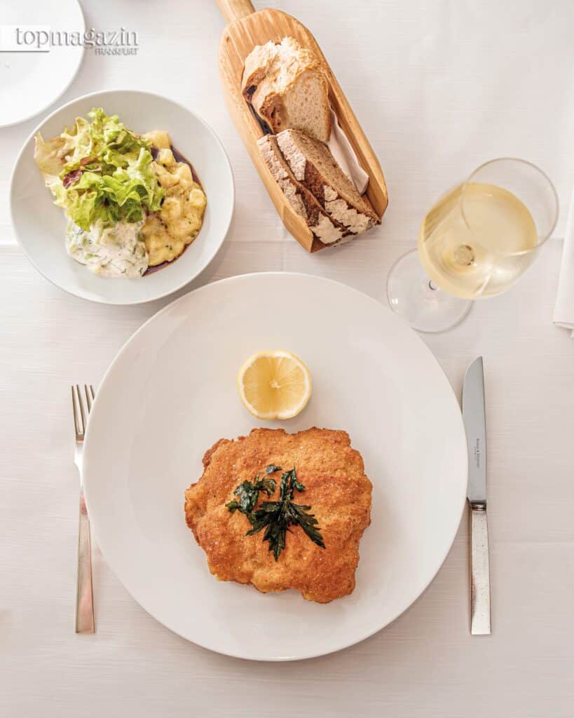 Ein Wiener Schnitzel von oben fotografiert, serviert mit einem leichten Salat und einem Glas Weißwein – ein kulinarischer Klassiker in Frankfurt.