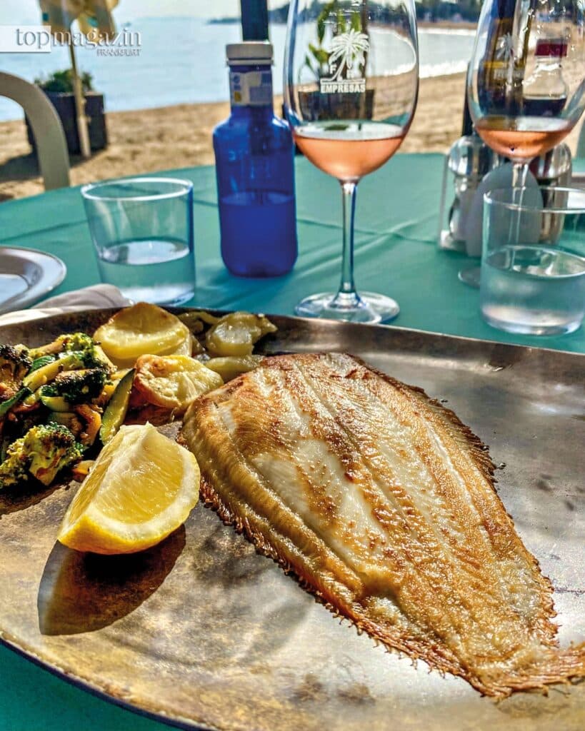 Eine perfekt gebratene ganze Seezunge auf einem silbernen Teller vor einem Weinglas, im Hintergrund der Strand.