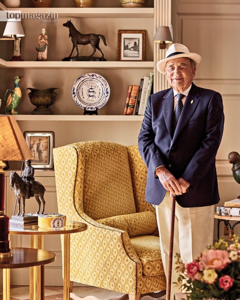 Conde Rudi im eleganten blauen Sakko, heller Hose und Strohhut stehend vor einem klassischen Interieur im Marbella Club.