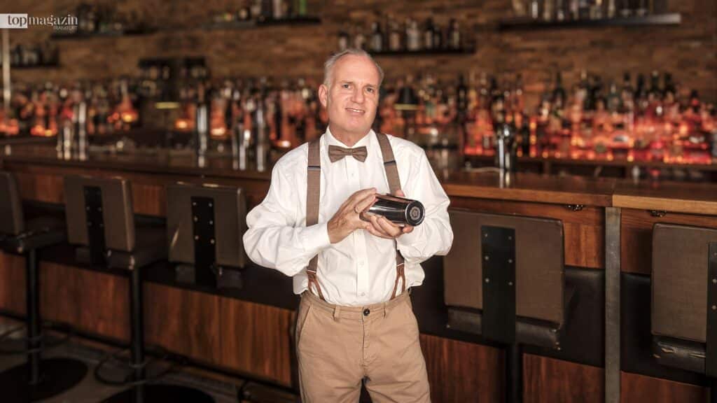 Der neue Barchef Emmanuel Saridakis steht mit weißem Hemd, Fliege und Hosenträgern lächelnd mit einem Cocktail-Shaker hinter dem Tresen in einer Bar.
