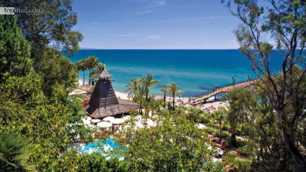 Der Eingang zur eleganten Strandbar MC Beach direkt am Meer mit weißen Sonnenschirmen, Sand und blauem Himmel.