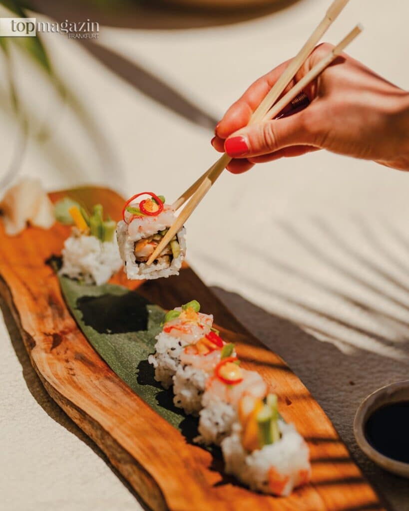 Eine Hand hält mit Essstäbchen ein kunstvoll angerichtetes Stück Sushi über einem rustikalen Holzteller in der Sonne.