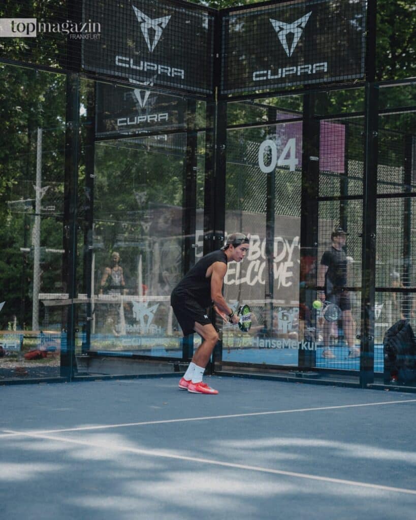 Ein Padel-Spieler schlägt den Ball auf einem Outdoor-Court nahe der gläsernen Rückwand.