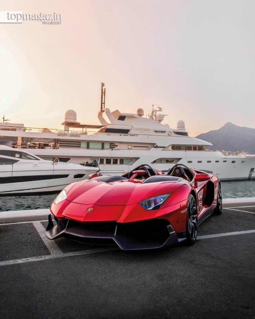 Ein roter Lamborghini parkt im Hafen von Puerto Banús direkt vor großen weißen Luxusyachten vor der Kulisse der Berge.
