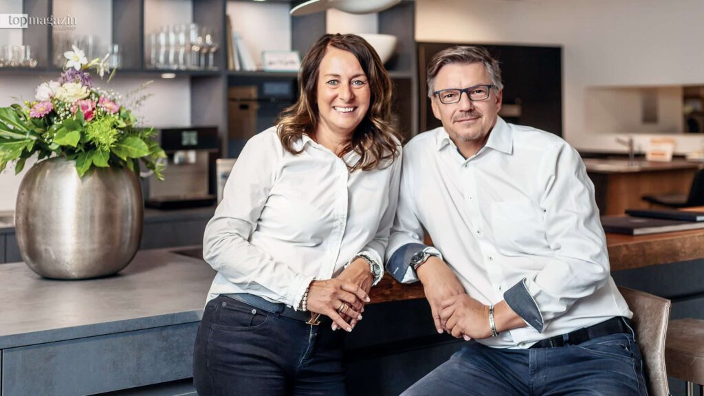 Sabine und Gerhard Grohs, Inhaber des Küchen Ateliers, lächeln sympathisch nebeneinander stehend in ihrem Küchen-Showroom in Eschborn.