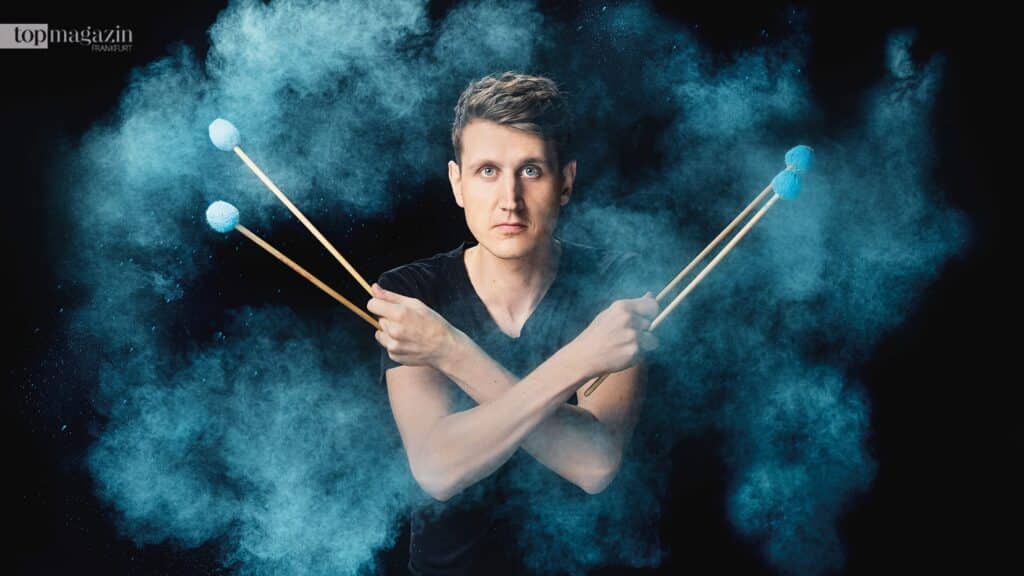 Der Perkussionist Alexej Gerassimez kreuzt zwei Trommelstöcke vor einer dynamischen, blauen Pulverwolke.