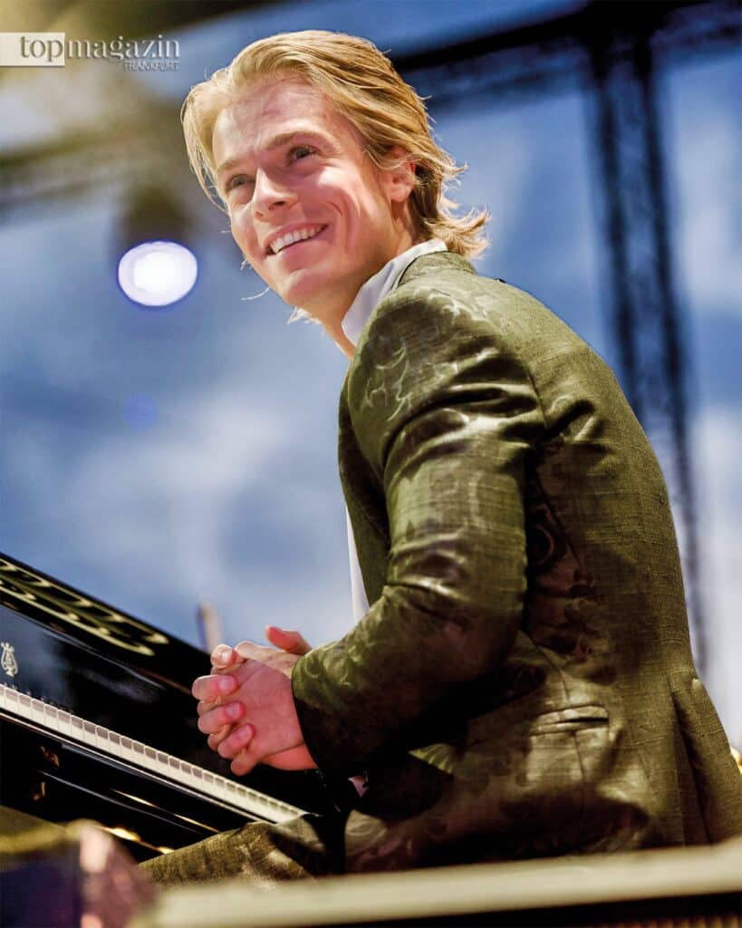 Der lachende niederländische Pianist Arthur Jussen sitzt am Flügel bei einem Open-Air-Konzert.