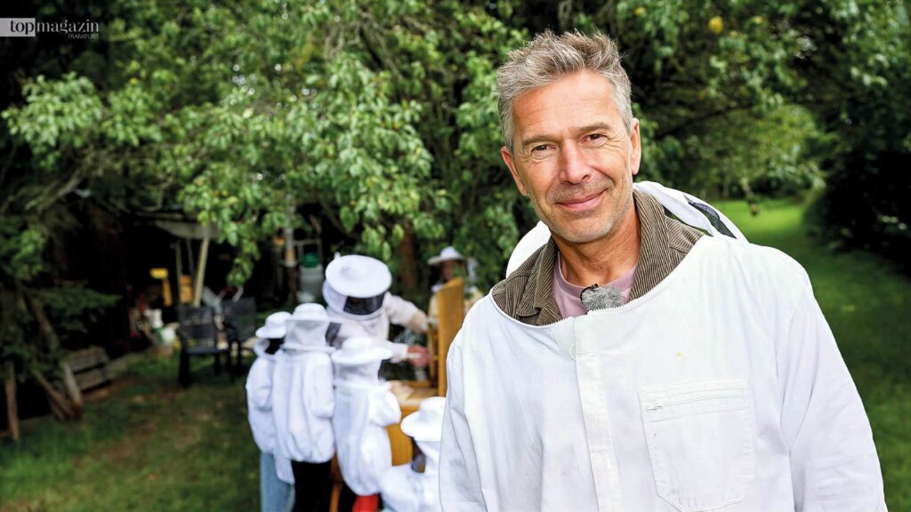 Dirk Steffens in weißer Imkerkleidung betrachtet mit Kindern in Schutzanzügen Bienenstöcke auf einer grünen Wiese.