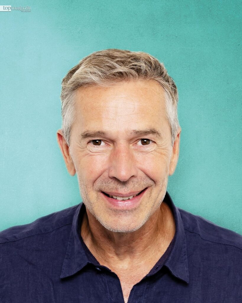 Porträt von Dirk Steffens, der freundlich und optimistisch vor einem hellblauen Hintergrund in die Kamera blickt.