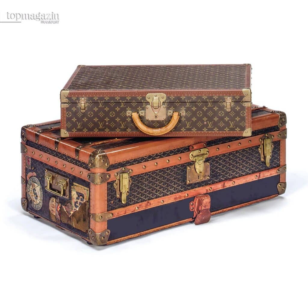 Zwei aufeinandergestapelte Vintage-Luxuskoffer von Louis Vuitton und Goyard vor weißem Hintergrund.