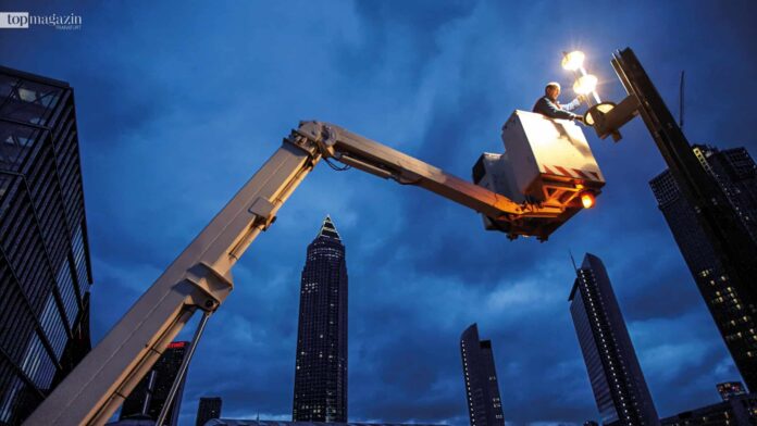 Techniker in einem Hubsteiger an einer Straßenlaterne, im Hintergrund der Frankfurter Messeturm in der Abenddämmerung
